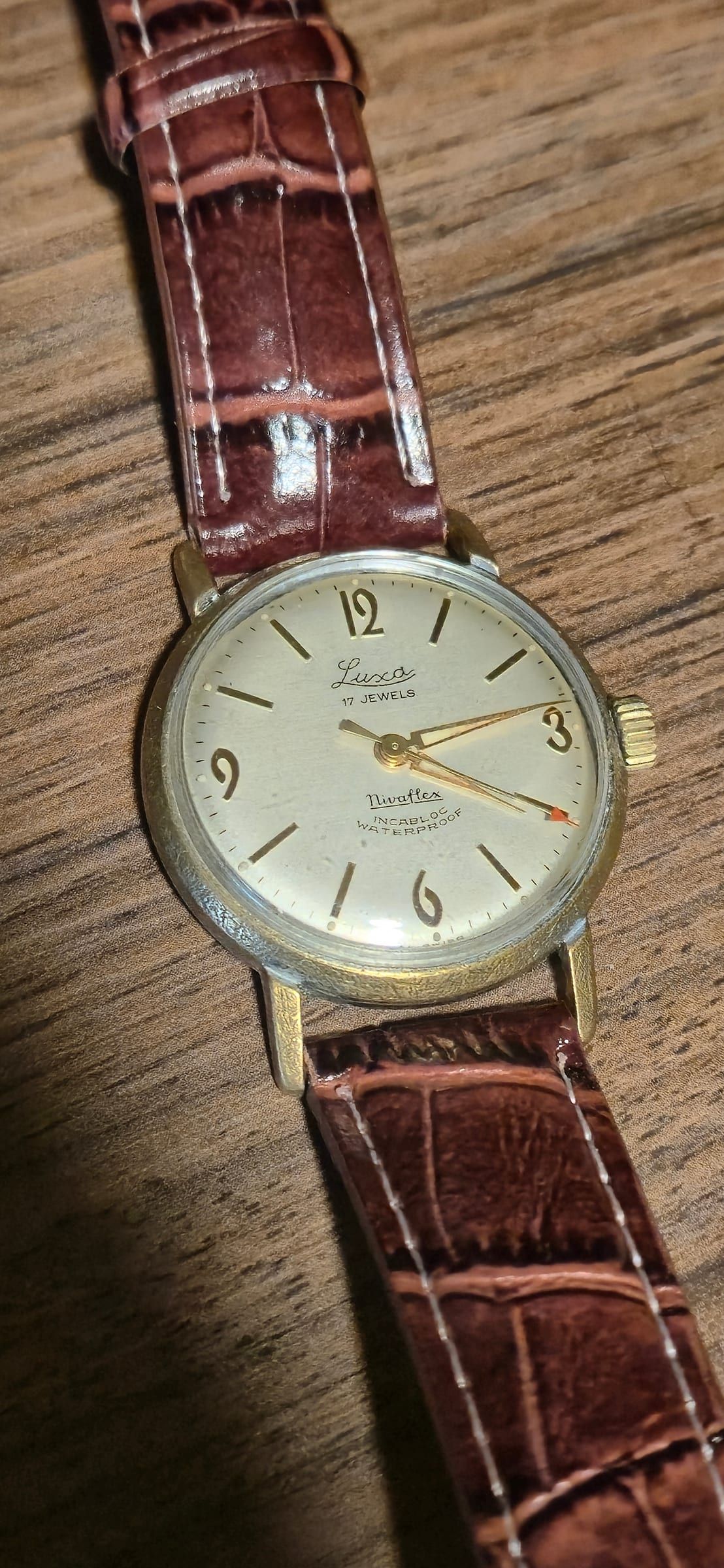Een vintage Luxa met crème wijzerplaat en gouden indexen, met '17 Jewels, Nivaflex, Incabloc, Waterproof', aan een bruine band.